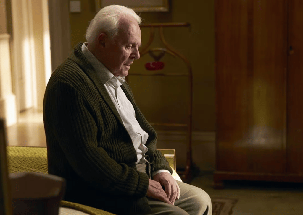 An elderly man in a green cardigan seated on a bench, looking contemplative with an expression of sadness.