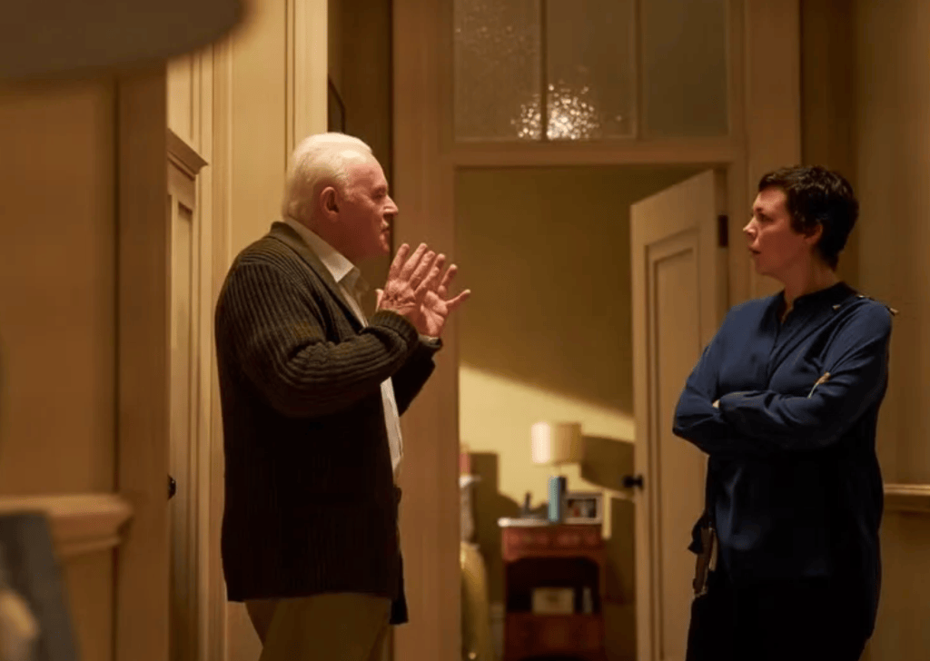 An elderly man in a dark sweater is animatedly speaking with a woman in a blue shirt, both standing in a well-lit hallway with an open door behind them.
